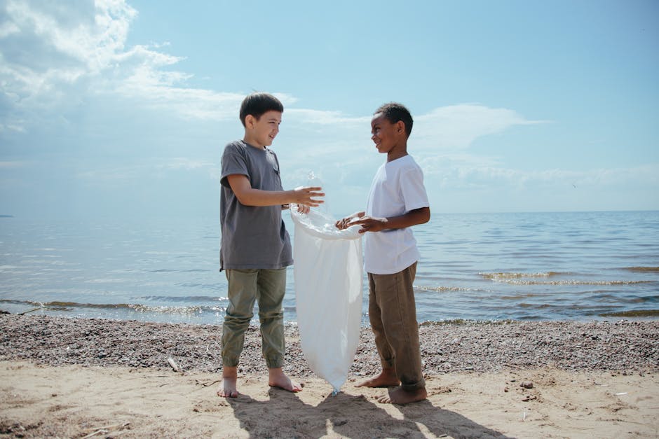Two young boys are standing on a sandy beach near the water, engaged in an activity involving a large white plastic bag. The boy on the left, wearing a grey t-shirt and light grey pants, is holding th
